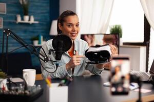 New media influencer smiling at camera while recording vr headphones review for followers. Creative vlogger making blog concept speaking and looking at smartphone on tripod home studio podcast photo
