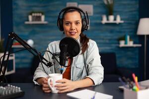Vlogger messaging her audience while making podcast in home studio for social media. Blogger recording online talk show using headphones, professional microphone looking at camera during broadcast. photo