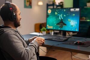 Pro man gamer playing game at professional computer late night wearing headphones. Excited player using wireless controller for virtual tournament gaming space shooter at home photo