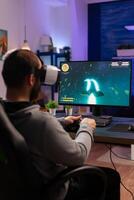 Player man playing game at powerful computer late night wearing vr headset. Excited player using wireless controller for virtual tournament gaming space shooter at home photo