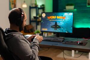 Back shot of pro gamer playing online shooter game on computer using wireless controller. Competitive player man winning videogame tournament use professional equipment at home gamming studio photo