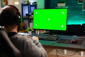Professional cyber gamer playing digital videogames on powerful computer with green screen display. Player using pc with mock up chroma isolated desktop streaming shooter games wearing headset photo