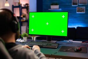 Professional player streaming videogames with green screen mock-up computer display in gaming home studio. Gamer using pc with chroma key isolated desktop streaming shooter games wearing headset photo