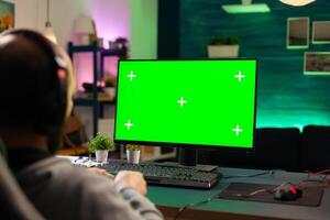 Concentrate gamer playing games on powerful computer with green mock up while streaming online competition. Player using pc with green screen isolated desktop streaming shooter games wearing headset photo