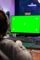 Gamer man playing videogames on computer with green screen holding wireless joystick in professional gaming studio. Player using pc with mock up isolated desktop streaming shooter games wearing headset photo