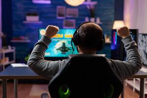 Back view of happy gamer playing and winning space shooter game in gaming home room. Cyber performing on powerful computer streaming videogames using professional headphones for online championship photo