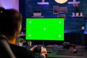 Pro gamer playing virtual videogames on professional powerful computer with green screen display. Cyber Player using pc with mock up chroma isolated desktop streaming shooter games wearing headset photo