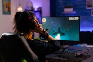 Focused Gamer puts professional headphones and playing space shooter videogames use modern equipment for digital tournament. Pro player sitting on gaming chair streaming games using RGB keyword photo