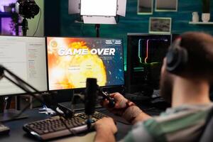 Back view of upseted streamer playing game competition using professional setup with streaming chat open. Gamer sitting on gaming chair using wireless controller and professional headphone photo