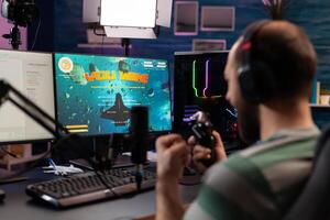 Over shoulder footage of professional streamer playing digital videogames on computer using headphones, microphone and controle. Streaming Man raising hands for wining space shooter game photo