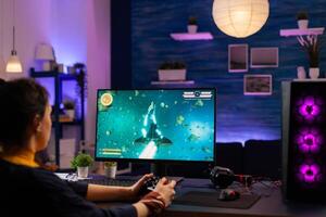 Concentrated player sitting on gaming desk late at night using wireless controller playing space shooter. Virtual streaming cyber performing live tournament using professional equipment photo