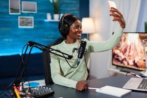 African influencer recording podcast and taking selfie in home studio. On-air online production internet podcast show host streaming live content, recording digital social media. photo