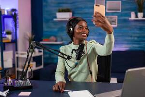Afro woman taking selfie with smartphone and using professional gear to record episode in living room. On-air online production internet podcast show host streaming live content, recording digital social media. photo