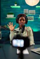 Afro content creator saying hellow to listeners wearing headphones. On-air production internet broadcast host streaming live content, recording digital social media photo