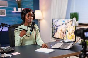 Afro blogger recording podcast talking into microphone in living room. Speaking during livestreaming, blogger discussing in podcast wearing headphones. photo