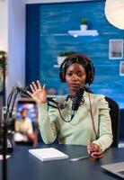 African blogger looking at camera and waving to camera recording episode for online show. Speaking during livestreaming, blogger discussing in podcast wearing headphones. photo