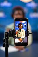 Close up of smartphone recording vlog of african influencer in home studio. Speaking during livestreaming, blogger discussing in podcast wearing headphones. photo