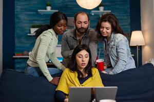 Happy friends having fun drinking beer talking on call using laptop. Group of multiracial people spending time together sitting on couch late at night in living room. photo