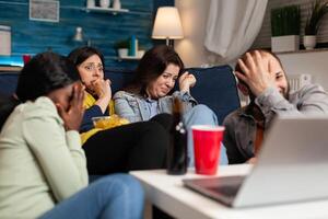 Multiracial friends chilling on sofa sitting in front of television watching horror thriller movie during night series. Group of multi-ethnic people having scary reaction, screaming with emotions. photo
