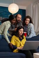 Multi ethnic friends hanging out talking on webcam laptop during call. Group of multiracial people spending time together sitting on couch late at night in living room. photo