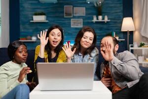 Happy multi-ethnic friends greeting her collegue during online videocall meeting using laptop webcam. Group of multiracial people spending time together on couch late at night during party photo