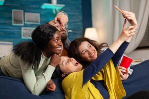 Cheerful diverse groupg of friends taking selfie photos having fun, drinking beer, sitting on couch socialising. Multhiethnic peple posting picturez on internet sharing with other person.
