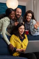Multi ethnic friends waving at laptop webcam during call sitting on couch late at night. Group of multiracial people spending time together sitting on couch late at night in living room. photo