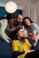 Multi ethnic friends bonding and talking on smartphone during call in home living room. Group of multiracial people spending time together sitting on couch late at night in living room. photo