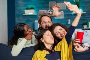 Multi-ethnic friends bonding together taking pictures making diverse expression posting on social media. Group of mixed race people laughting while sitting on sofa late at night in living room. photo