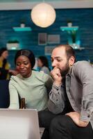 Multi ethnic friends socializing celebrating friendship watching funny on laptop sitting on couch. In background two women drinking beer enjoying time spend together during entertainment party. photo