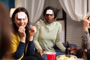 Group of multi-ethnic people having fun while playing board game using sticky notes spending time together. Creative friends hanging out late at night in living room during home party. photo