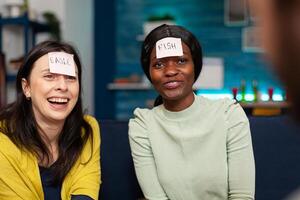 Multiracial friends enjoying friendship while playing guess who game attaching sticky notes on forehead. Mixed race people having fun, laughing together while sitting on sofa late at night. photo