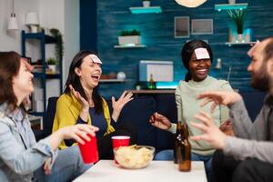Mixed race friends playing board game using sticky notes spending time together late at night relaxing on couch in living room. Group of multi-ethnic people drinking beer, eating chips enjoying home party. photo