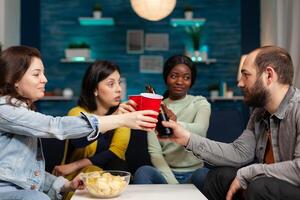 Happy cheerful multi ethnic friends clinking beer while having fun and entertaining themselfGroup of multiracial people spending time together sitting on couch late at night in living room. photo