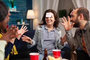 Mixed race friends socializing during entertainment party while playing guess who game attaching stickey notes. Multiracial people spending time together having fun during entertainment wekeend. photo