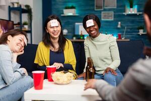Mixed race friends playing Guess who game with sticky papers attached on forehead. Group of multiracial people having fun, laughing together while sitting on sofa in living room late at night. photo