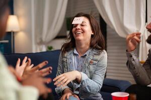 Multiethnic friends having attaching sticky notes on forehead while playing guess who game. Mixed race people having fun, laughing together while sitting on sofa in living room late at night. photo