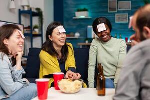 Mixed race friends gathering in living room playing guess who game using sticky notes. Mixed race people having fun, laughing together while sitting on sofa late at night during funny party. photo