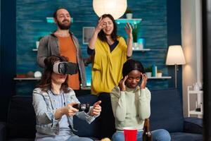 Multiethnic friends experiencing virtual reality losing game wearing vr headset during gaming competition. Mixed race group of people hanging out together having fun late at night in living room. photo