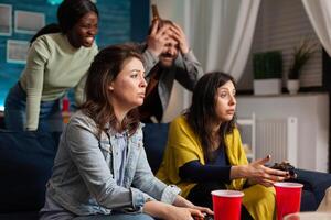 Multi-ethnic friends sad after losing games competition while socialising using wireless controller sitting on couch, drinking beer late at night on big tv. photo