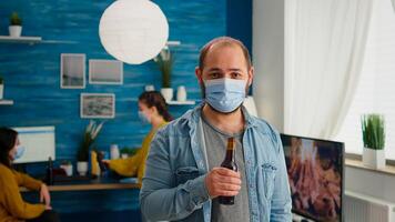 Man with protection mask looking at camera holding bottle of beer ready for party with friends in living room respecting social distance in global pandemic. People socializing during covid 19 outbreak photo
