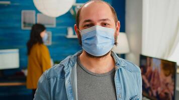 Portrait of man putting on protection mask looking at camera spending good time with friends in living room respecting social distance in global pandemic. People socializing during covid 19 outbreak photo