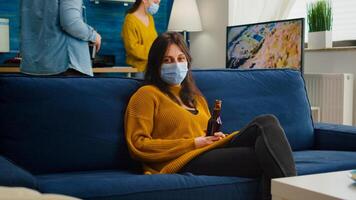 Woman relaxing with friends sitting on couch wearing face mask against spreading coronavirus during global pandemic holding beer bottle looking at camera. People socializing during covid 19 outbreak photo