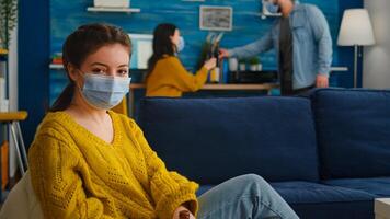 Lady looking at camera enoying spending time with friends in living room keeping social distancing wearing face mask to prevent coronavirus spreading. during global outbreak holding beer bottle. photo