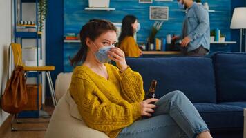 Woman holding beer bottle drinking looking at camera, relaxing on sofa, spending time with friends keeping social distancing with face mask in living room preventing spread of coronavirus. photo