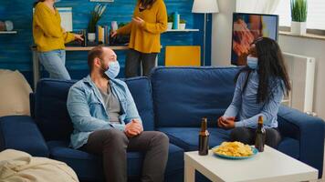 Diverse friends discussing during new normal party wearing protection mask, socializing keeping distance sitting on sofa drinking beer and eating snaks. Group of multi ethnic people enjoying free time photo