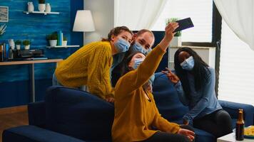 Multiracial friends taking selfie with face masks during covid 19 outbreak, new normal lifestyle concept with people having fun in living room respecting social distance to prevent virus spread photo