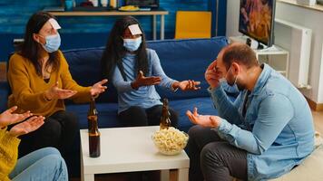 Thoughtful multiethnic friends with sticky notes on foreheads playing name game miming gesturing wearing face mask keeping social distancing preventing covid19 spread. People enjoying bottles of beer photo