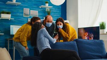African woman showing funny clip on smartphone hanging out in home living room wearing face mask preventing coronavirus during global pandemic keeping social distancing, enjoying party in outbreak photo