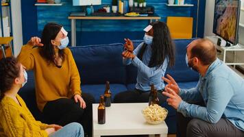 Happy cheerful multi ethnic friends with face mask playing games with sticky notes on forheads drinking beer eating popcorn guesing keeping social distancing. during outbreak in home living room photo